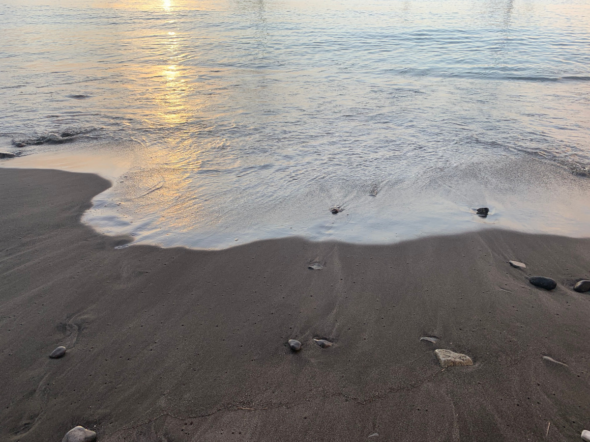 La lumière du soleil se reflète dans la vague sur la sable. Sa couleur irisée contraste avec l'aspect sombre du sable serti de roches polies.  