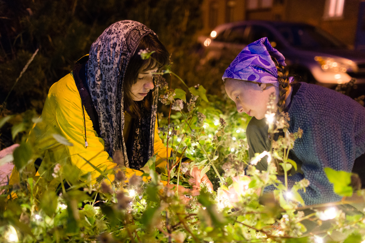 Deux jeunes personnes sont accroupies dans un bosquet aux fruits lumineux. Leurs cheveux sont couverts de foulards. Elles semblent très attentives à ce qui se trouve au sol, entre elles. 