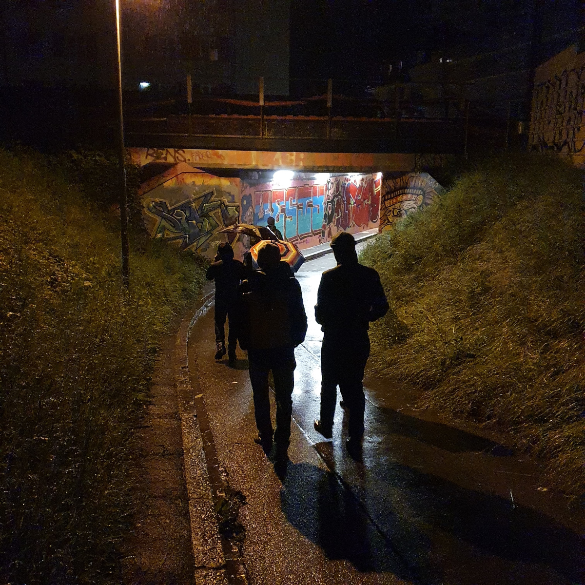 De nuit, un groupe portant imperméables et parapluies se déplace sous un viaduc coloré de graffitis.