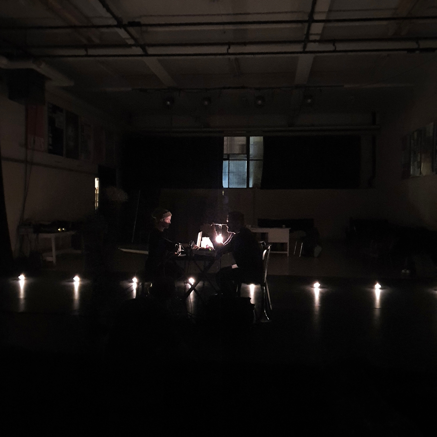 Deux personnes se font face dans un éclairage minimal, une rangée de lumières traversant la photo. Au fond, une fenêtre.