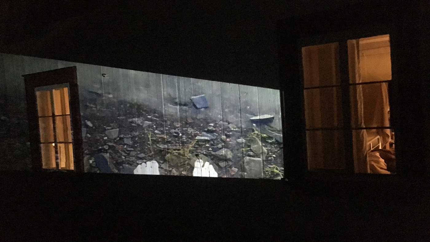 Projection d'une image de pieds et de fleuve sur le mur extérieur d'une maison, la nuit. À gauche, une porte entrouverte; à droite, une fenêtre.