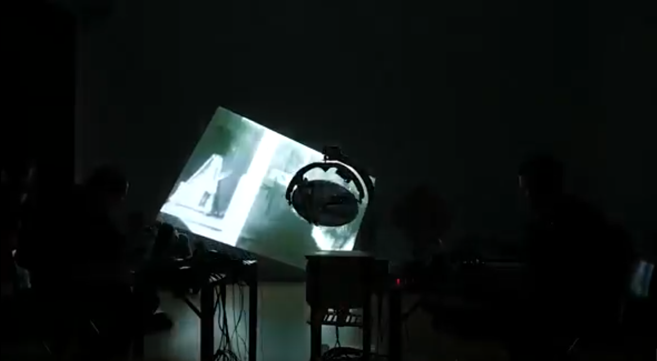 Dans la pénombre, on perçoit une projection lumineuse non orthogonale sur un mur et devant laquelle on devine un appareillage technologique.