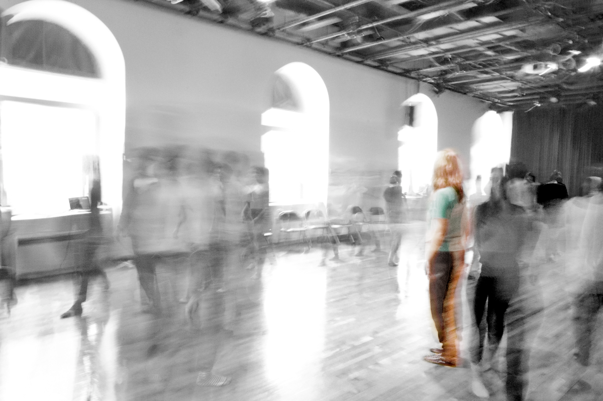 Une grande pièce avec de grandes fenêtres semble en noir et blanc et quelques personnes aux vêtements colorés s'y trouvent, rendues floues par le mouvement.