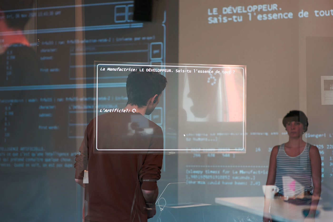 Deux interprètes relaient (et jouent) en temps réel la production de texte de la machine, à l’aide d’un système à l’oreillette. En surimpression&nbsp;: la réplique générée par la machine